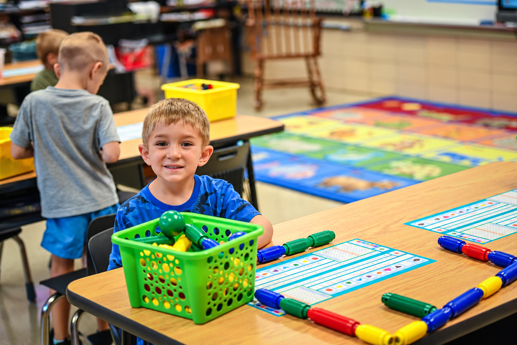 Children seated at desks. One child has a basket of colorful plastic rings and beads. Behind them, rugs on the floor.