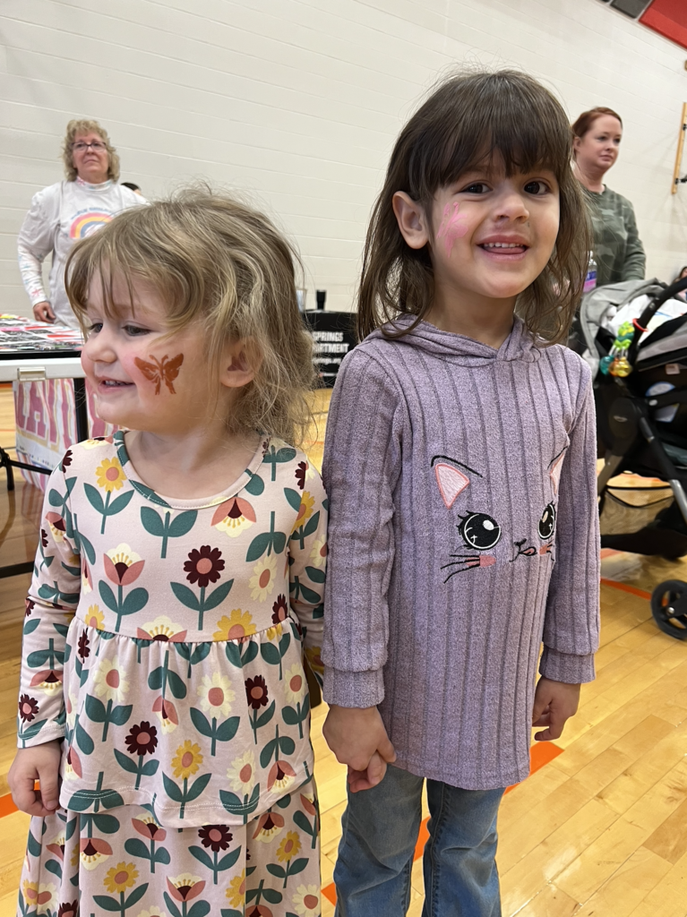 Two young girls smile and hold hands, both with face paint on their cheeks, standing in a brightly lit gym filled with carnival booths.