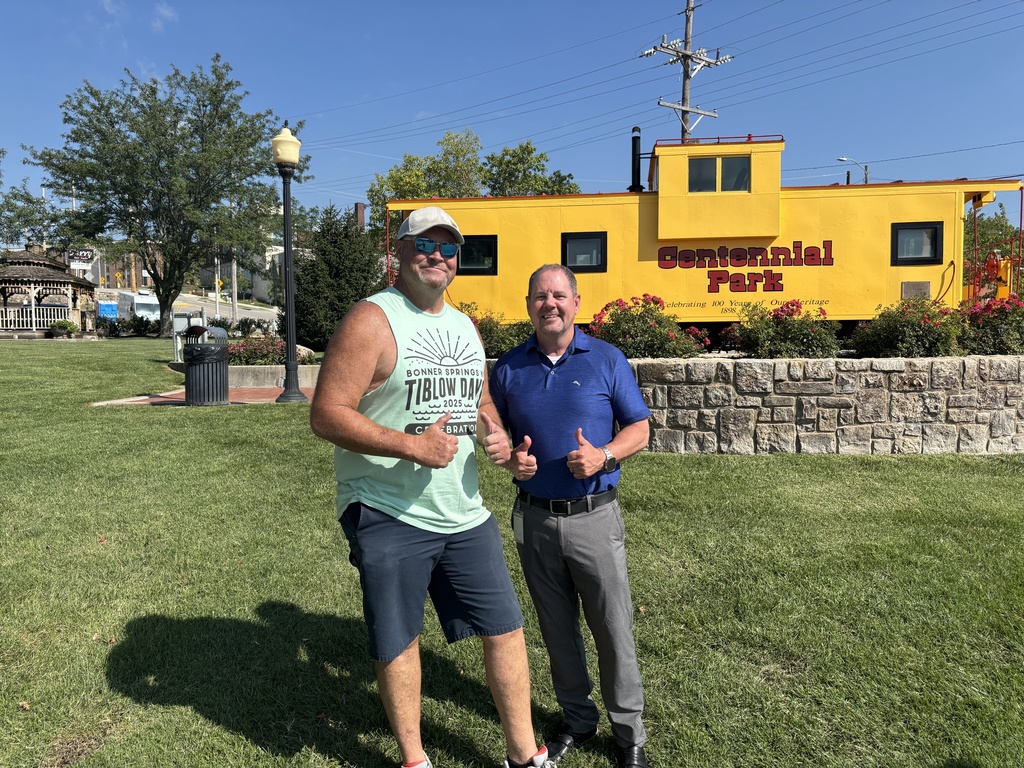 Two men smile and give thumbs up while standing on the grass in front of a bright yellow train car with the words “Centennial Park” painted on it. The man on the left wears sunglasses, a cap, shorts, and a sleeveless Tiblow Days shirt, while the man on the right wears a blue polo and slacks. Trees, a lamp post, and stone landscaping surround the scene on a sunny day.