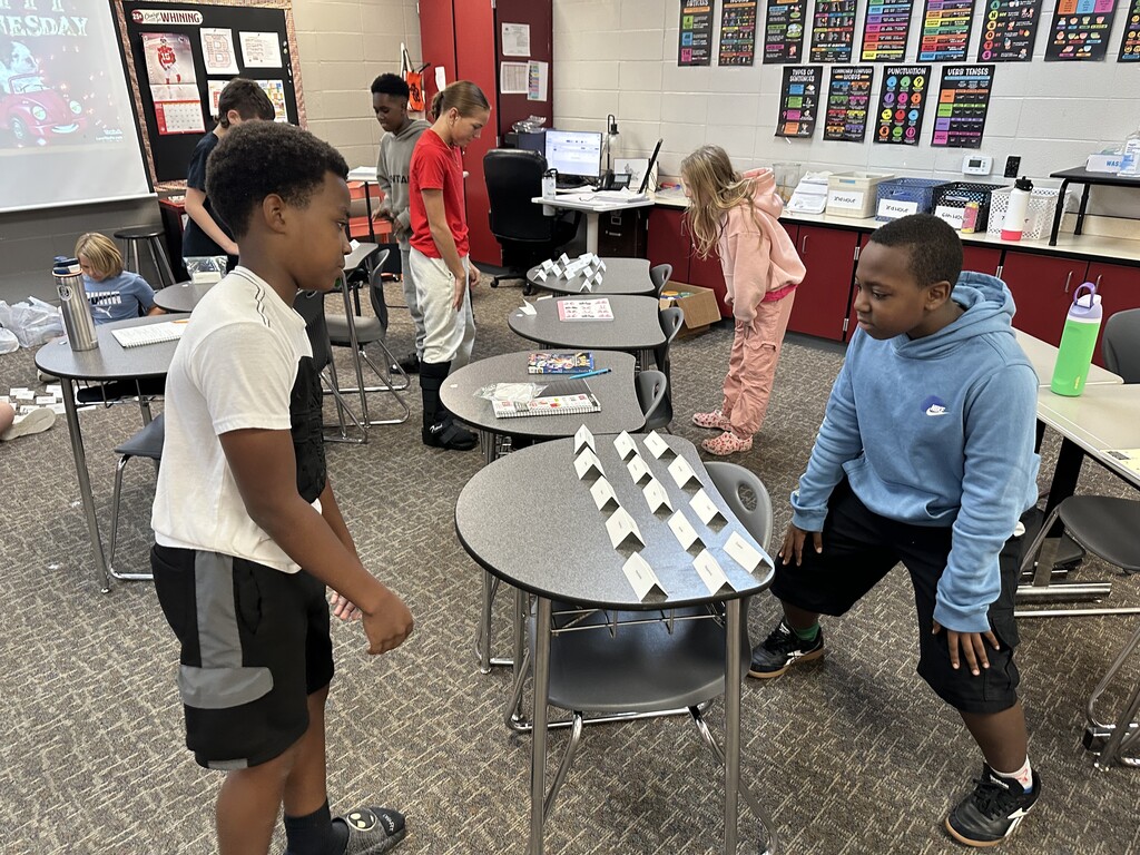 Two boys stand face-to-face at desks with vocabulary cards propped up, while other classmates work in the background.