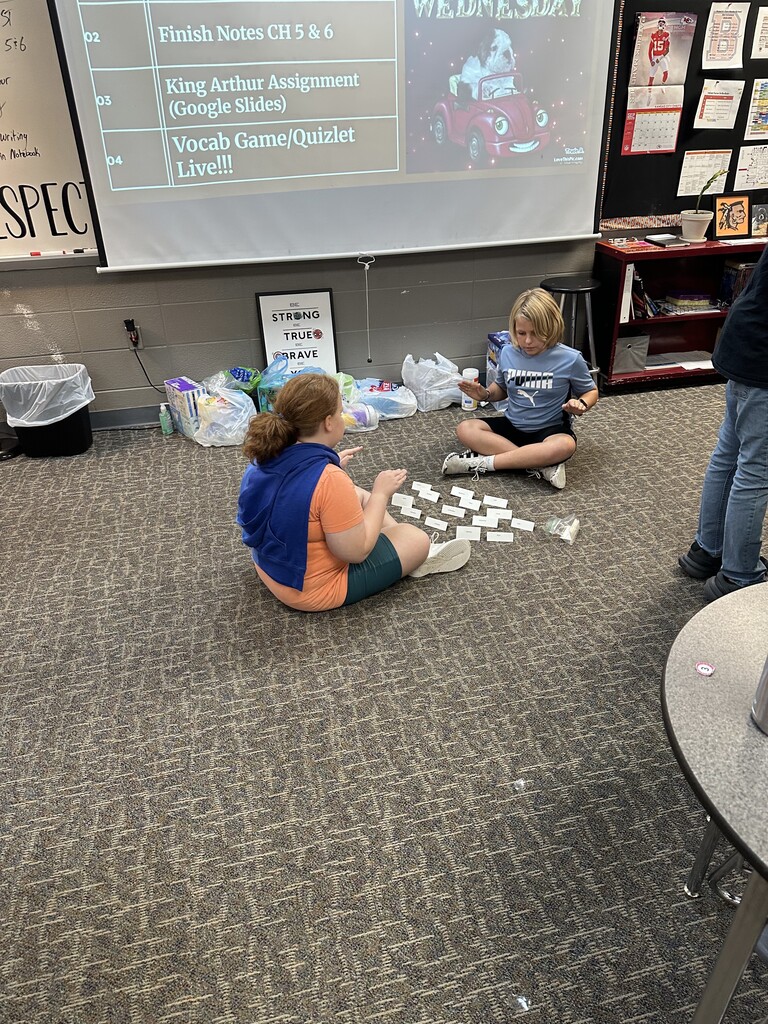 Two students sit cross-legged on the floor in front of a projector screen, sorting through vocabulary cards spread out in front of them.
