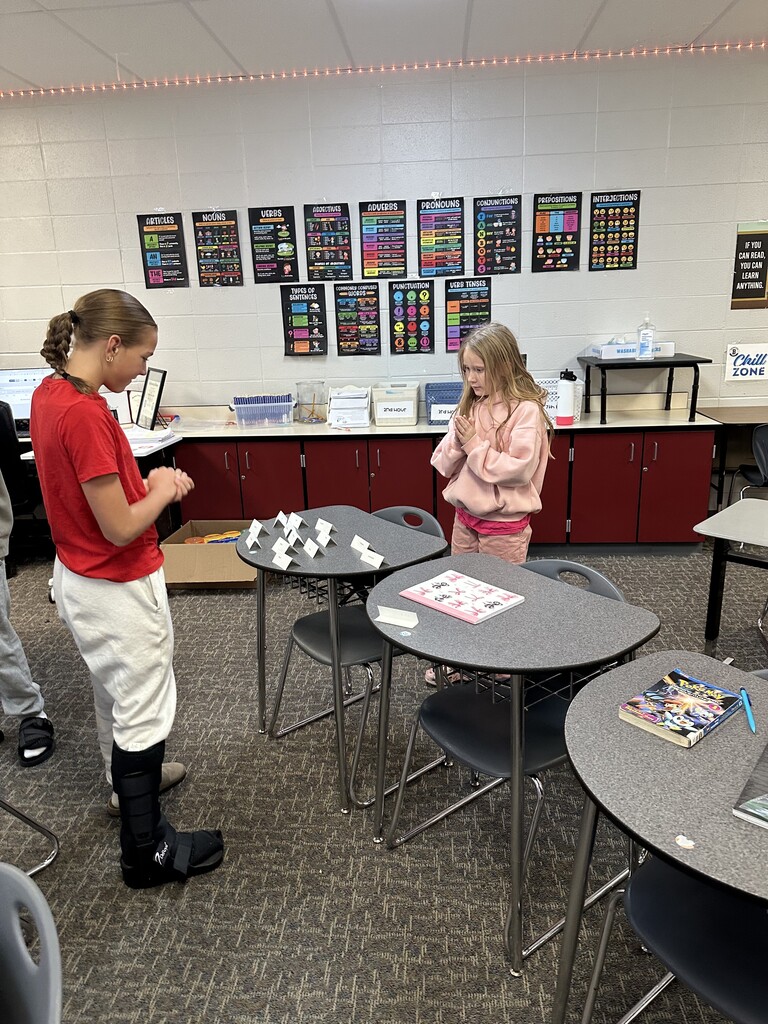 Two girls stand across from each other at desks with word cards set up, appearing to play a matching game.