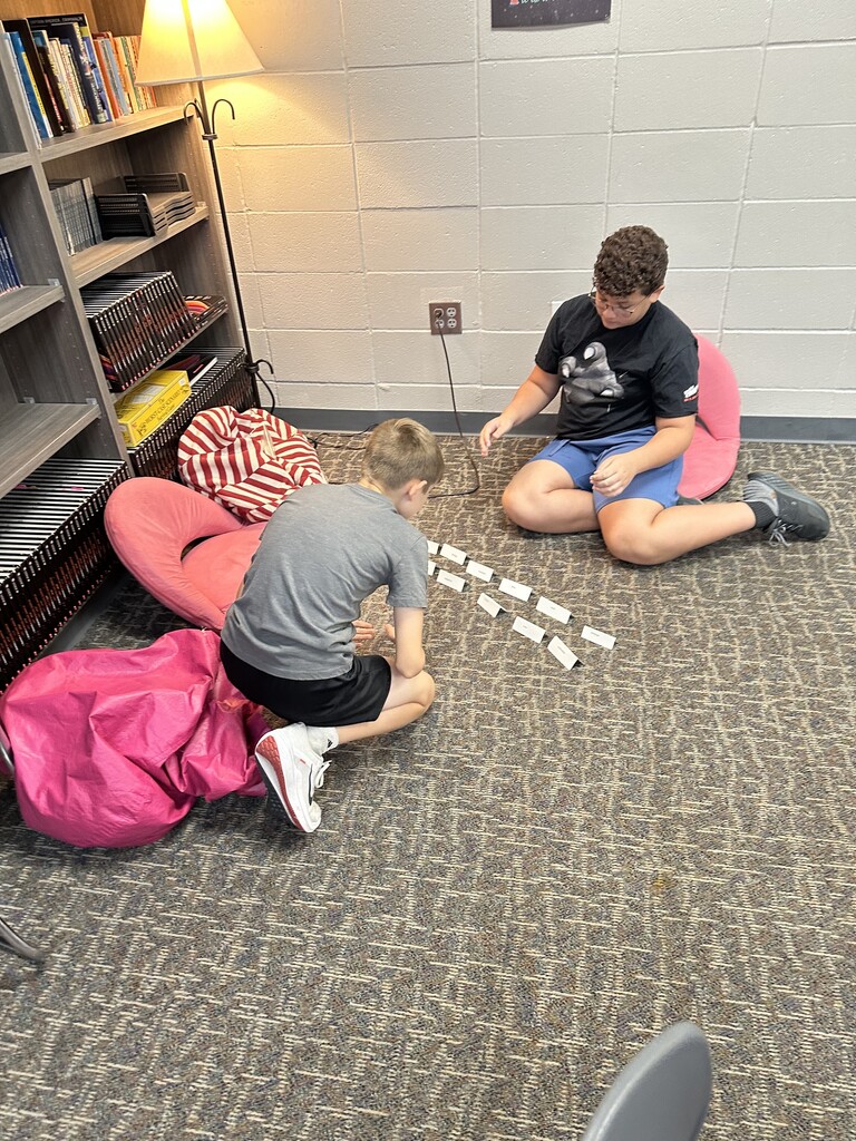 Two boys sit on the floor in a classroom corner with beanbag chairs and a lamp beside bookshelves. They are arranging vocabulary word cards in rows.