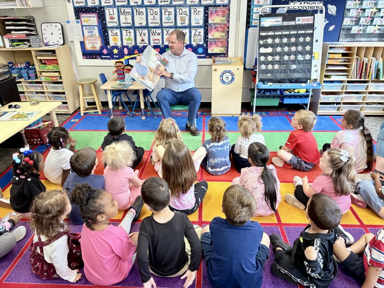 superintendent Moulin reading a book about dinosaurs to the class. students look on attentively.