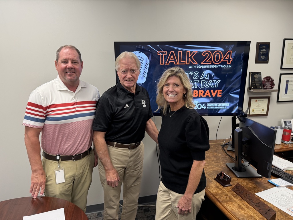 Three people stand together smiling in an office in front of a large screen displaying the Talk 204 podcast graphic that reads “It’s a great day to be Brave.” The person on the left wears a white and red striped polo, the person in the center wears a black polo with khaki pants, and the person on the right wears a black top and khaki pants. Framed awards and certificates decorate the wall, and a wooden desk with a computer and papers is visible in the foreground.
