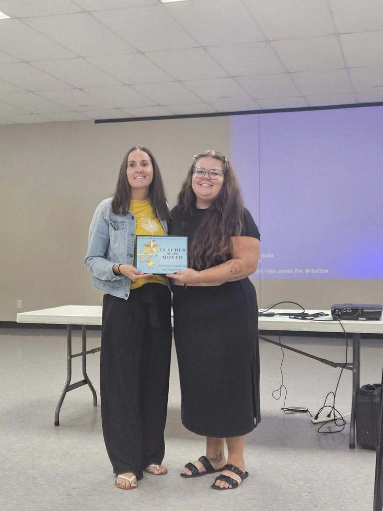 Two women stand together in a meeting room, smiling at the camera. One of them is holding a plaque that reads “Teacher of the Month.” They are standing in front of a table and projection screen during a Chamber of Commerce luncheon event.