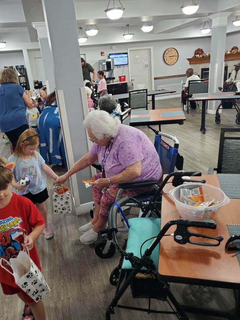 Children hand out treat bags to senior residents seated in a dining area. One elderly woman in a wheelchair reaches out to accept a bag from a child.