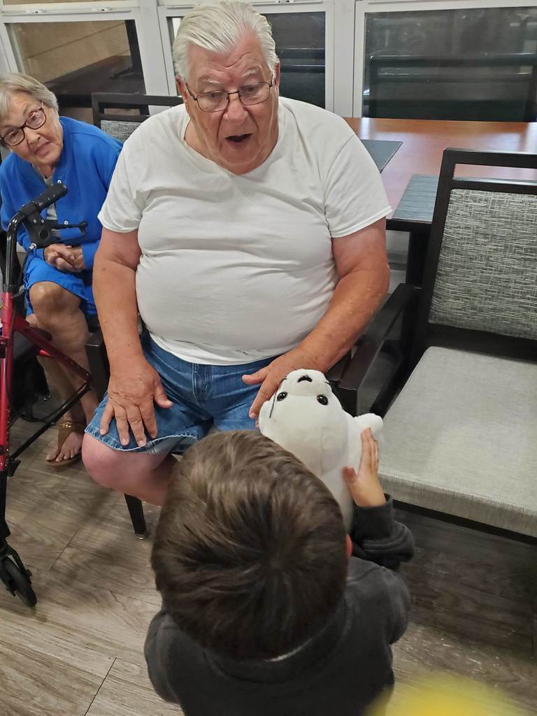 An older man sitting in a chair looks surprised and delighted as a young child shows him a white stuffed animal. An older woman sits nearby watching with a smile.