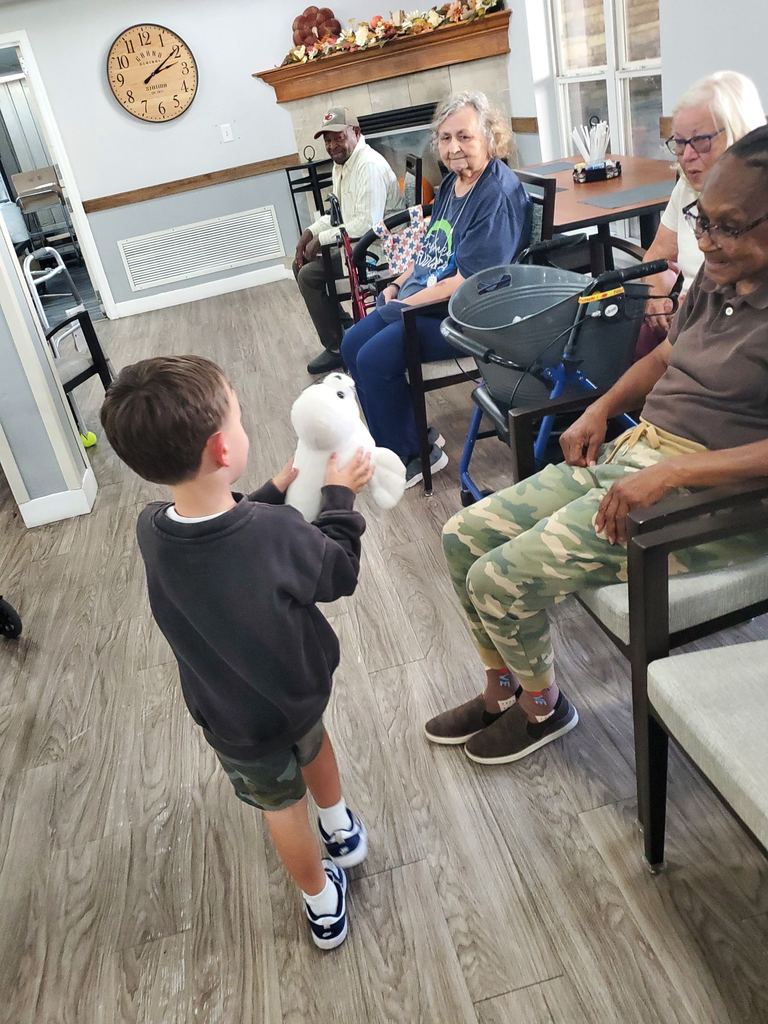 A young boy holds out a white stuffed animal to an older woman seated in a community room, while other residents look on and smile.