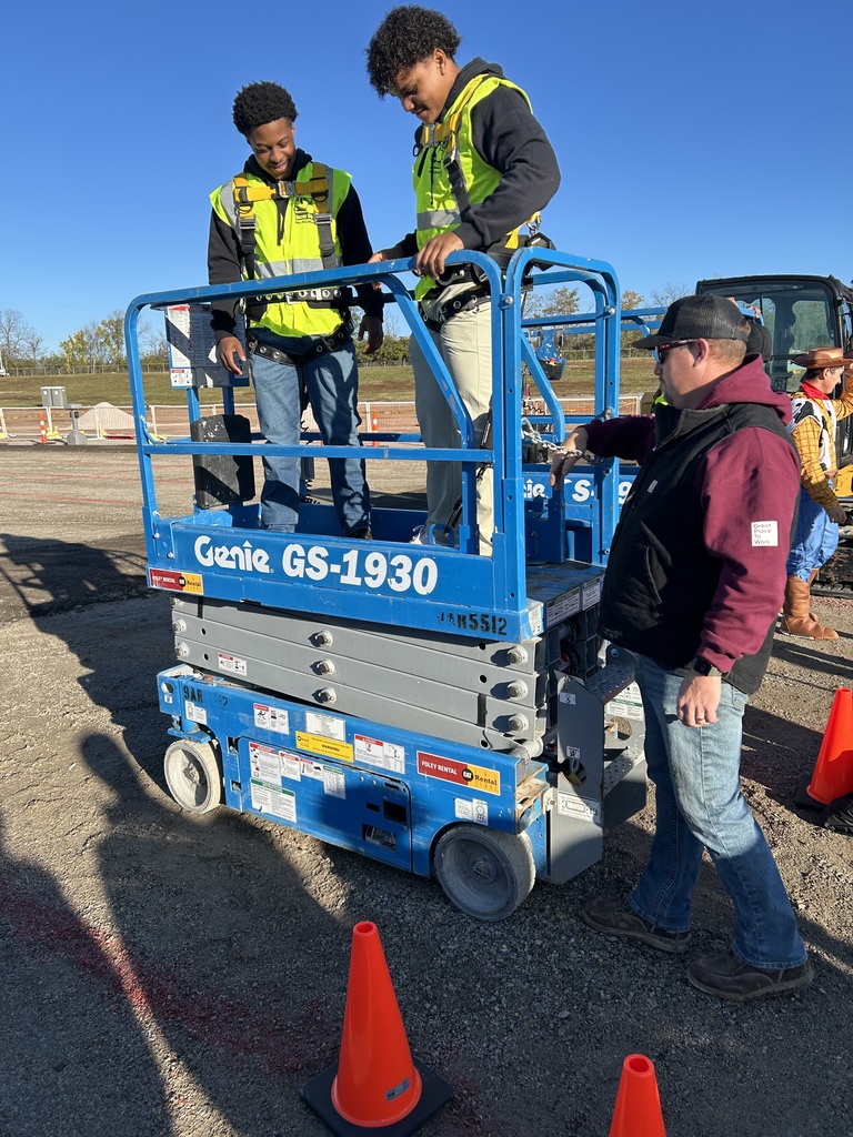 Two Bonner Springs High School students stand on a blue Genie GS-1930 lift, fastening safety harnesses under the supervision of an instructor at Dozer Day®. They are outdoors on a sunny day, surrounded by orange cones and construction equipment.
