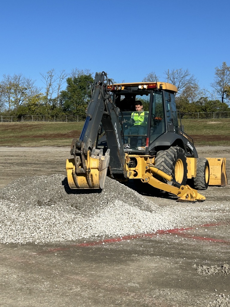 A Bonner Springs High School student operates a yellow backhoe loader, scooping gravel at Dozer Day®. The scene takes place on a large outdoor lot with piles of gravel and a bright blue sky in the background.