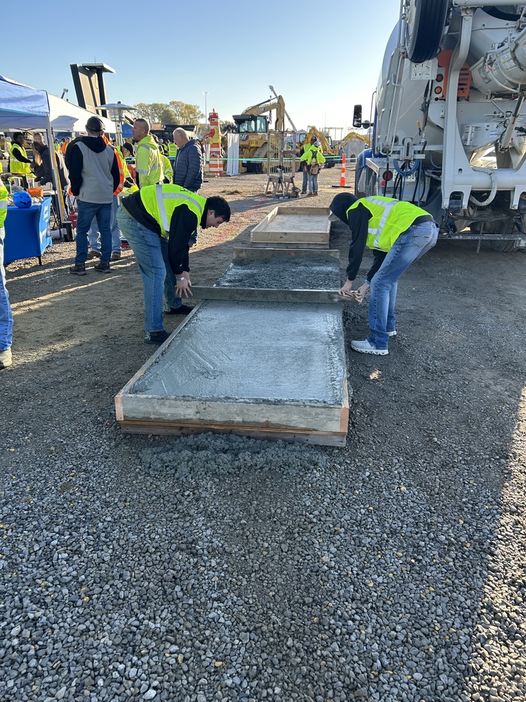 Bonner Springs High School students wearing bright yellow safety vests smooth a large section of freshly poured concrete at Dozer Day®. A concrete truck is parked beside them, and event tents, equipment, and other participants can be seen in the background.
