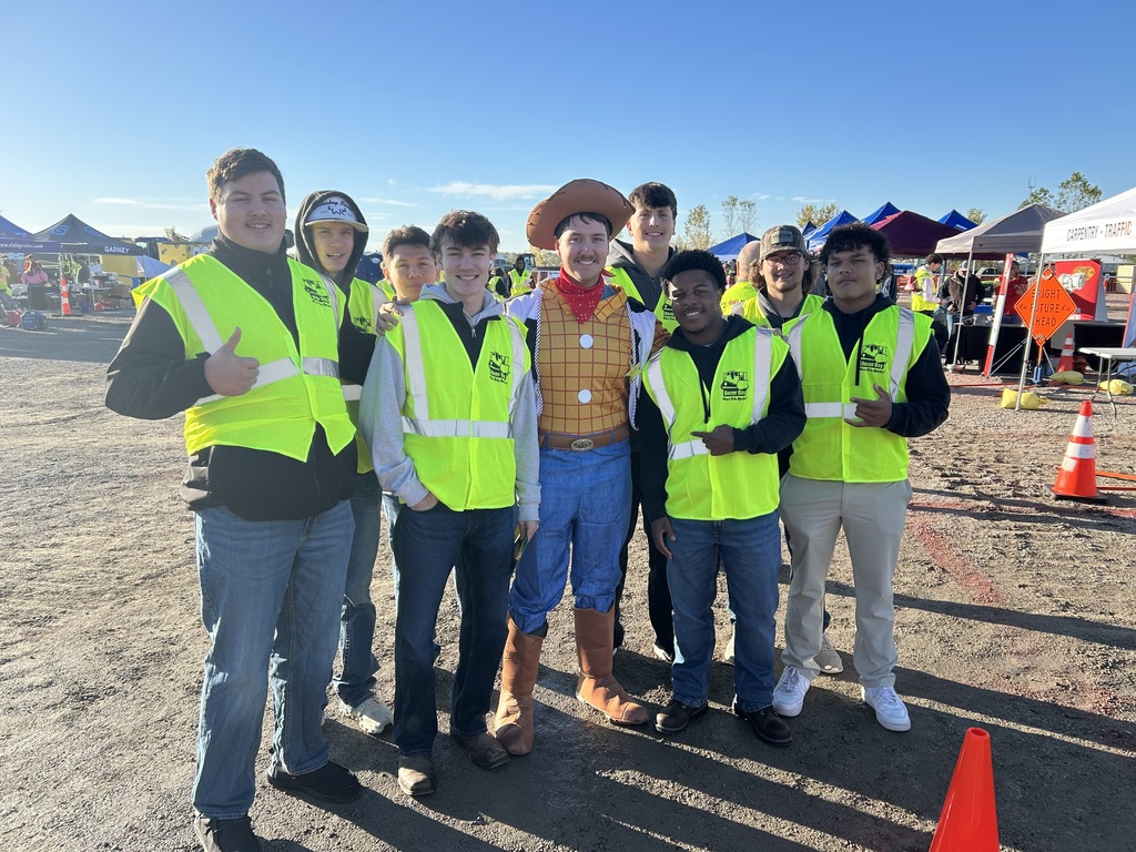 A group of Bonner Springs High School students pose for a photo with a person dressed as Woody from Toy Story at Dozer Day®. Everyone is wearing bright yellow safety vests, smiling, and standing under a clear blue sky surrounded by event booths and construction equipment.