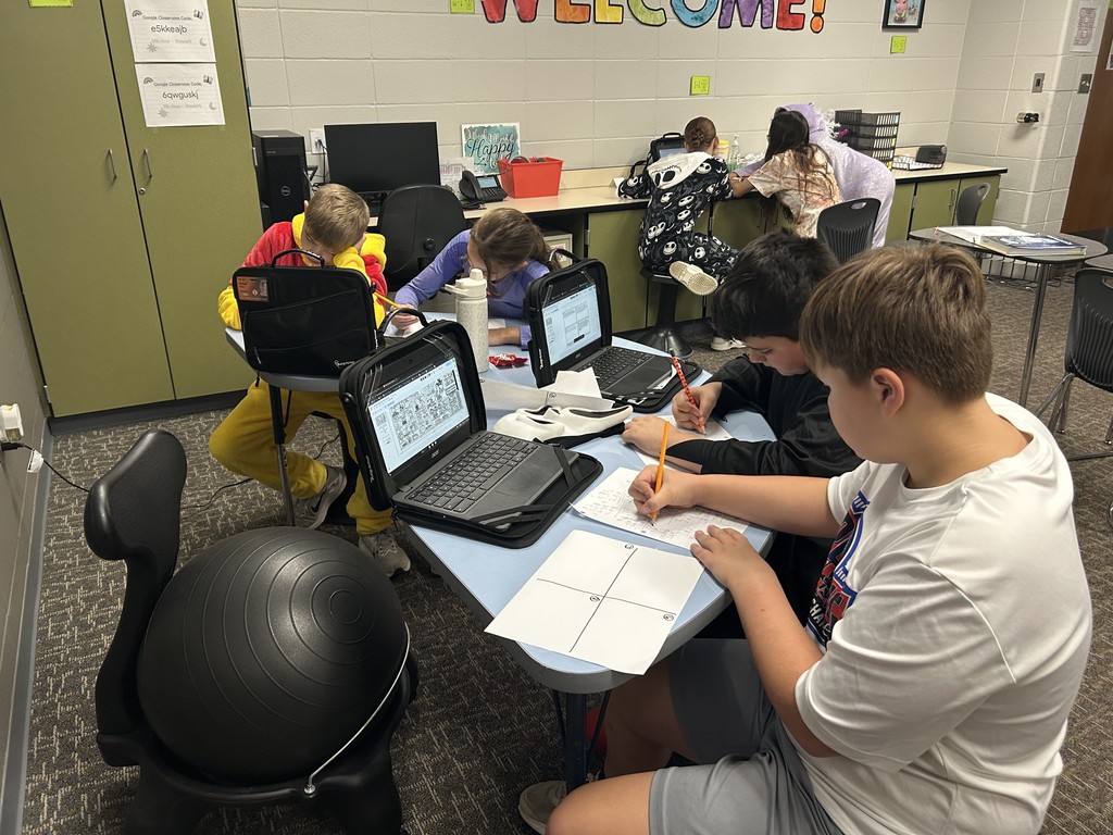 Several students, some dressed in Halloween costumes, work in groups on laptops and paper activities in a classroom. They’re collaborating on math problems at round tables.