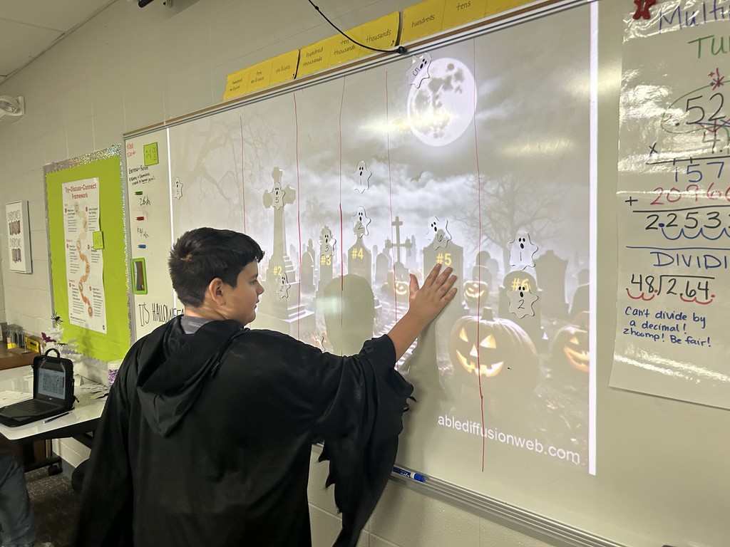A student dressed in a costume stands at the whiteboard, working on a Halloween-themed math activity projected with ghosts, pumpkins, and gravestones. The board includes numbers and math symbols related to decimal operations.