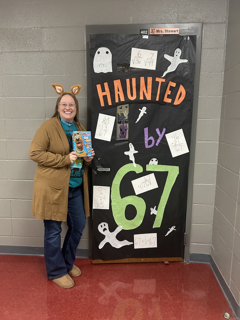A smiling teacher dressed in a Scooby-Doo-themed outfit stands beside a classroom door decorated with a Halloween theme. The door reads “Haunted by 67” with ghosts and math equations attached.