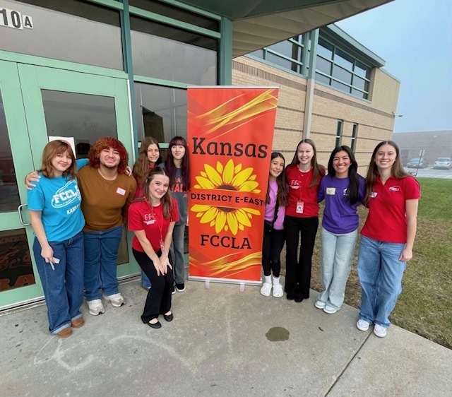 A group of FCCLA members from multiple schools stand outside next to a Kansas District E-East FCCLA banner, smiling in front of the school entrance.