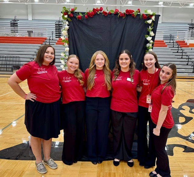 Six FCCLA members wearing matching red shirts pose together in front of a flower-decorated black backdrop in the gym.