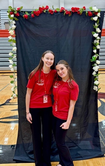 Two Bonner Springs High School FCCLA members smile while standing together in front of a black backdrop decorated with red and white roses inside the gym.