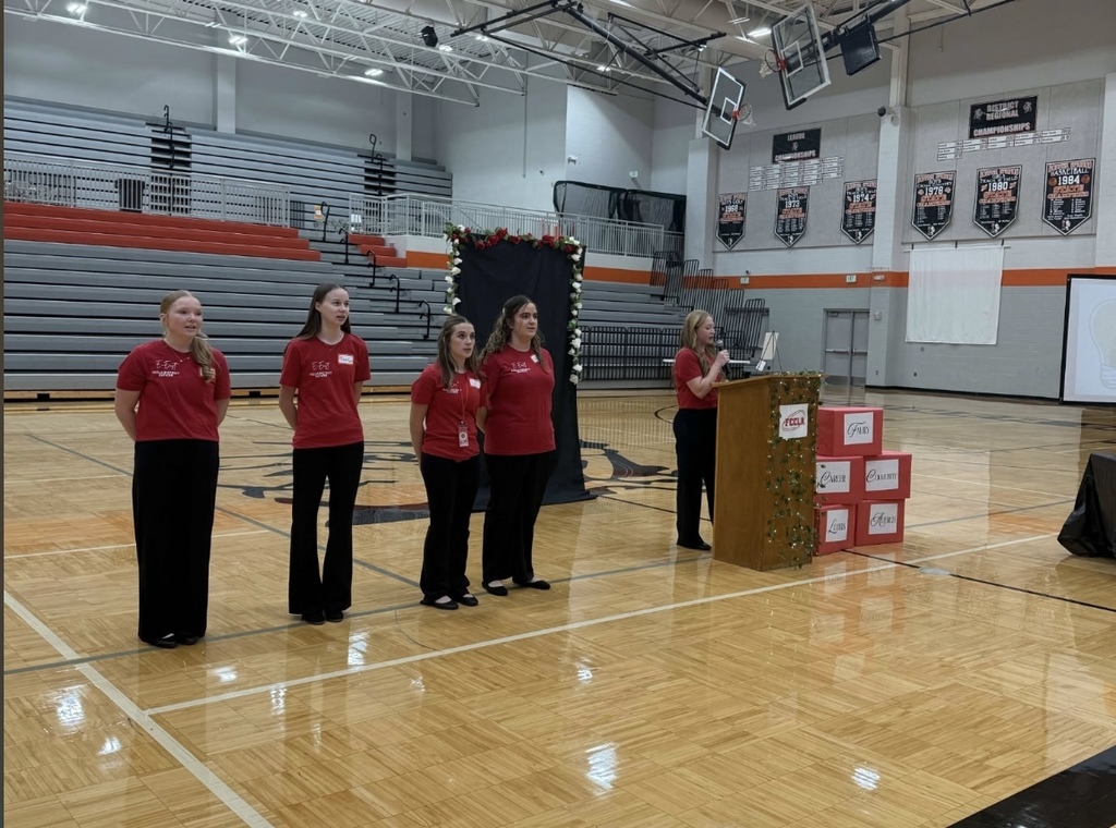FCCLA advisors and student leaders stand on the gym floor near a decorated podium, speaking and presenting to attendees during the Fall Leadership Conference.