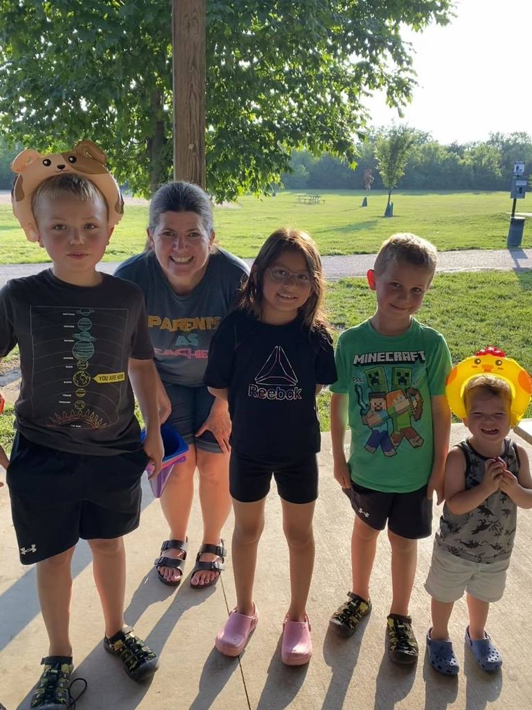 A woman wearing a ‘Parents as Teachers’ shirt stands under a park pavilion with four smiling children on a sunny summer day.