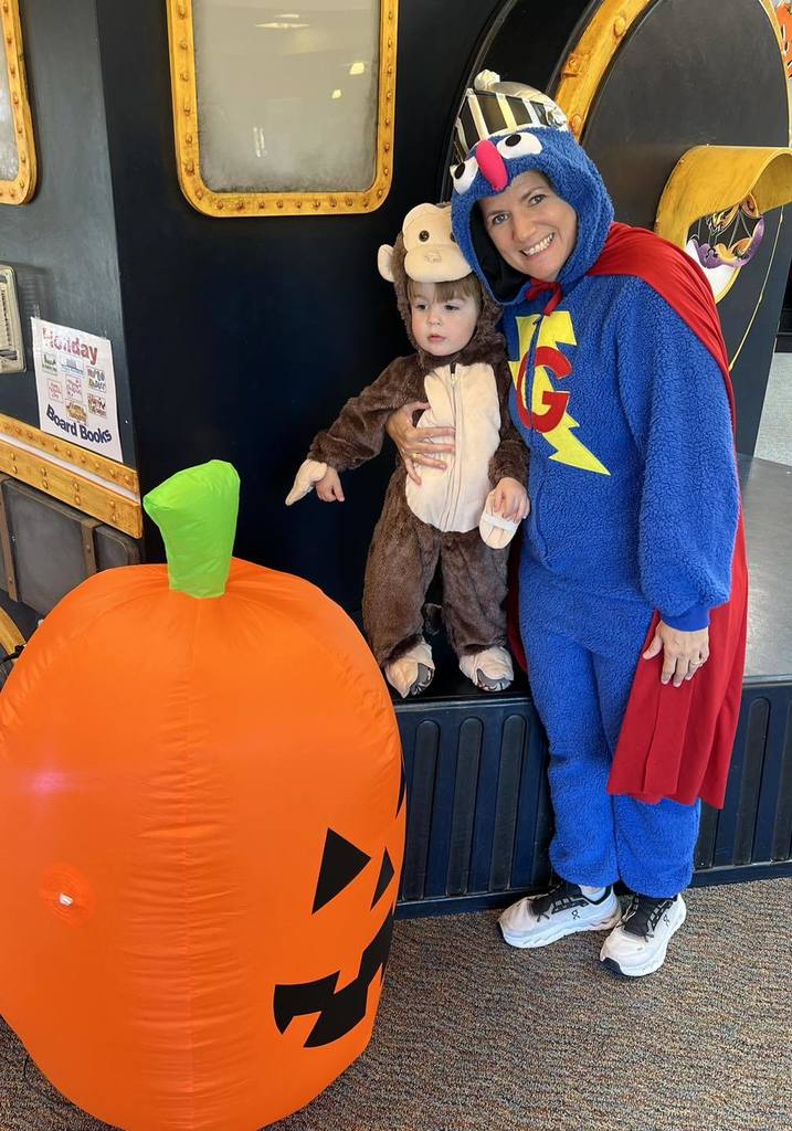 A woman in a blue superhero costume with a red cape poses with a toddler dressed as a monkey beside a large inflatable orange pumpkin at a Halloween event.