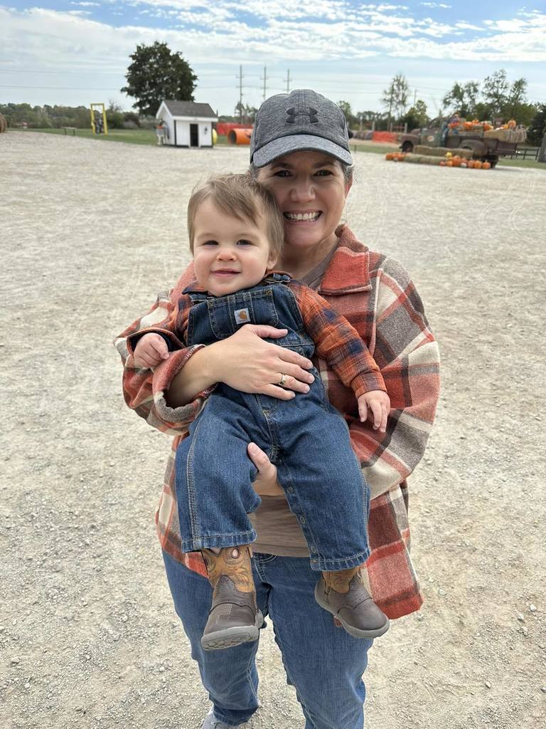 A smiling woman wearing a plaid jacket and gray cap holds a happy toddler dressed in overalls and cowboy boots at an outdoor pumpkin patch.