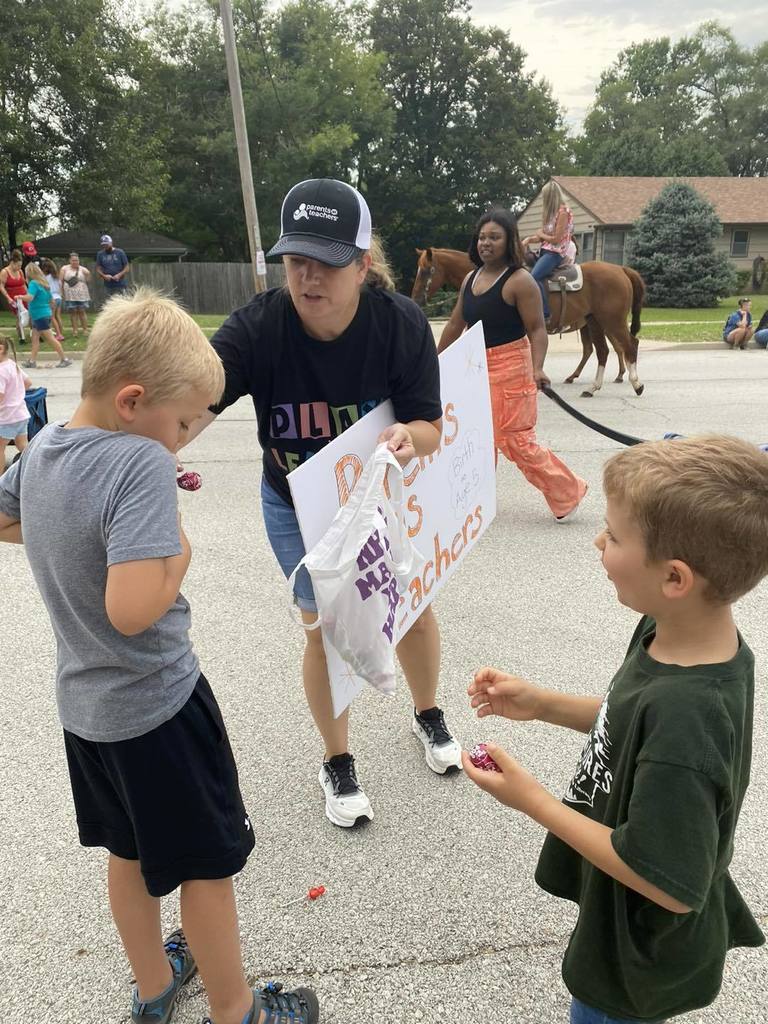 A woman wearing a black hat and ‘Parents as Teachers’ T-shirt hands out candy to children along a small-town parade route while families and a person on horseback watch in the background.