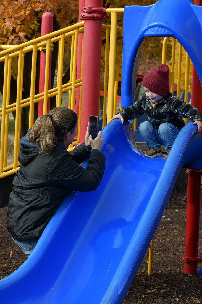 A woman kneels at the bottom of a playground slide taking a photo of a young child in a red hat and plaid jacket who is smiling at the top of the slide on a fall day.
