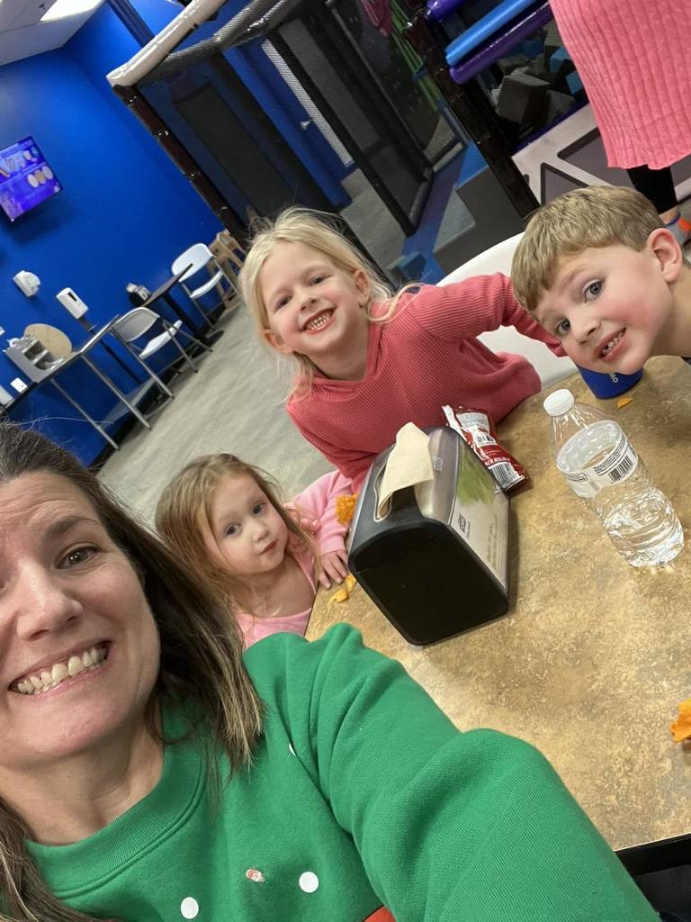 A woman in a green sweatshirt smiles for a selfie with three young children sitting at a table inside a brightly colored indoor play area.