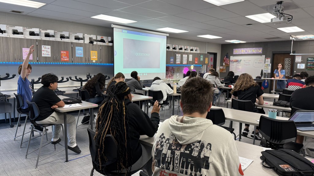 Students sit in a classroom at Clark Middle School listening to Meteorologist Stevie Stephenson from KCTV5 News give a presentation on meteorology. A slide reading “Questions?” is projected at the front as students raise their hands to participate.