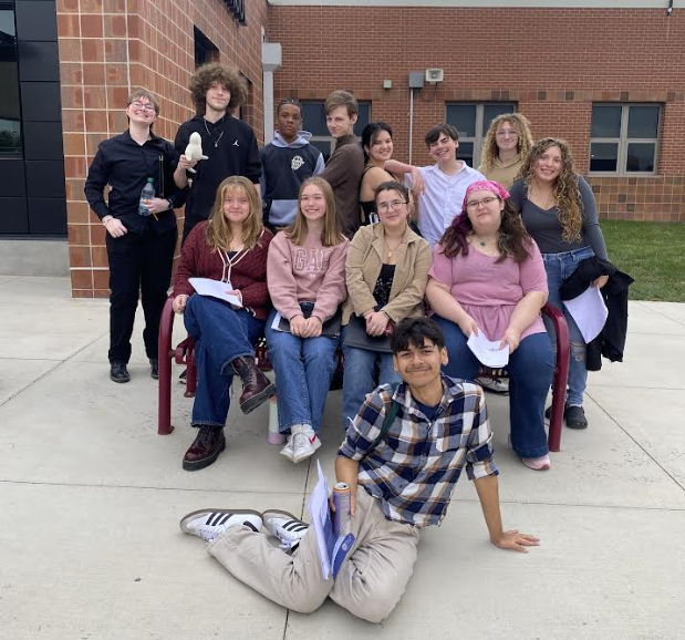 Bonner Springs High School choir students gathered outside near a brick building. The group is smiling, with some sitting on benches and others standing behind them. One student is sitting on the ground in front holding music papers.