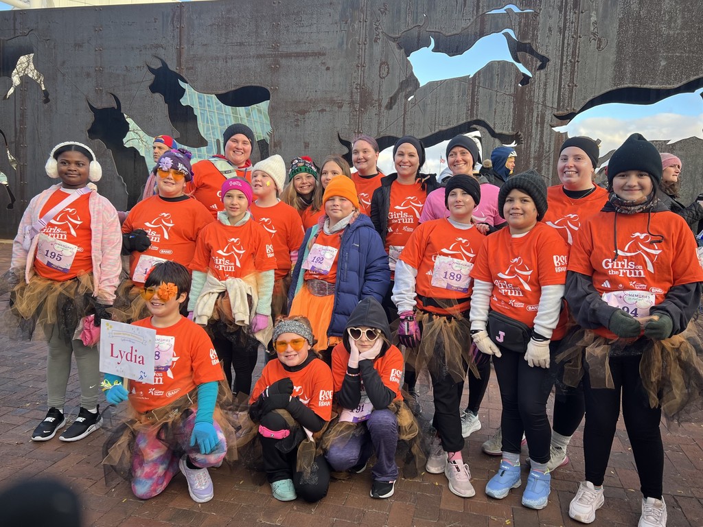 A group of girls and coaches from Bonner Springs Elementary pose together outdoors in bright orange Girls on the Run shirts. Many of the girls wear tutus, winter hats, earmuffs, and gloves. They are smiling and standing in front of a large metal art wall with animal silhouettes cut out. Race bibs are visible on several of the girls.