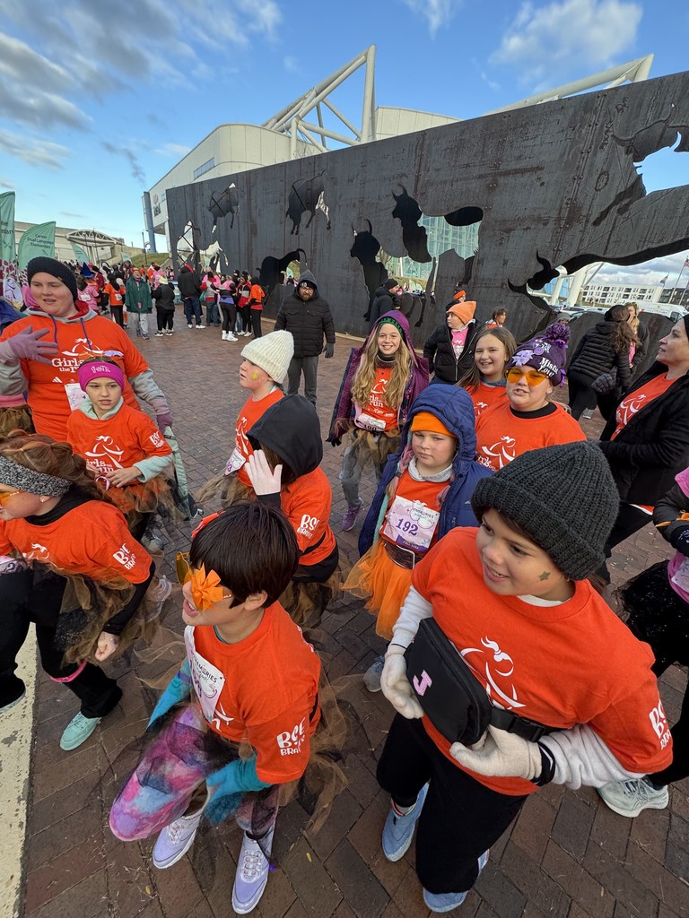 A candid shot of the Girls on the Run team gathered near the starting area. The girls wear orange shirts and winter clothing, some with face paint and tutus. They appear excited and energetic as people gather around them. The same large metal silhouette wall is visible in the background.