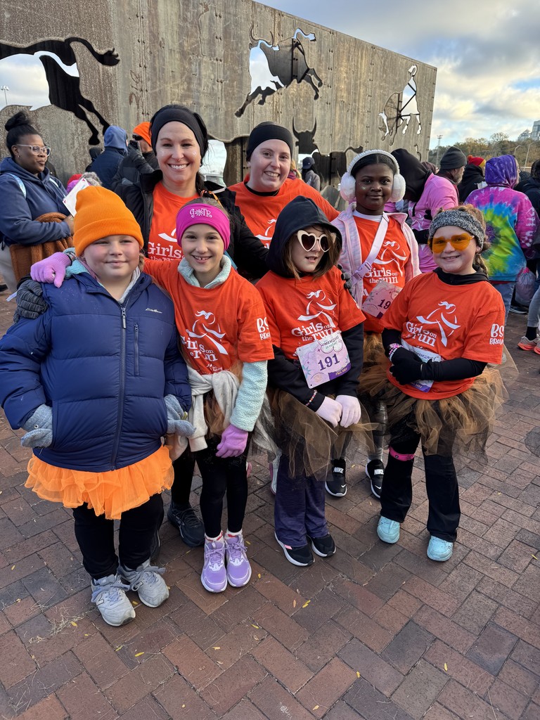A small group of girls and two coaches pose closely together, smiling at the camera. They are wearing bright orange Girls on the Run shirts along with winter hats, gloves, and tutus. The metal animal-silhouette wall and other race participants are visible in the background.