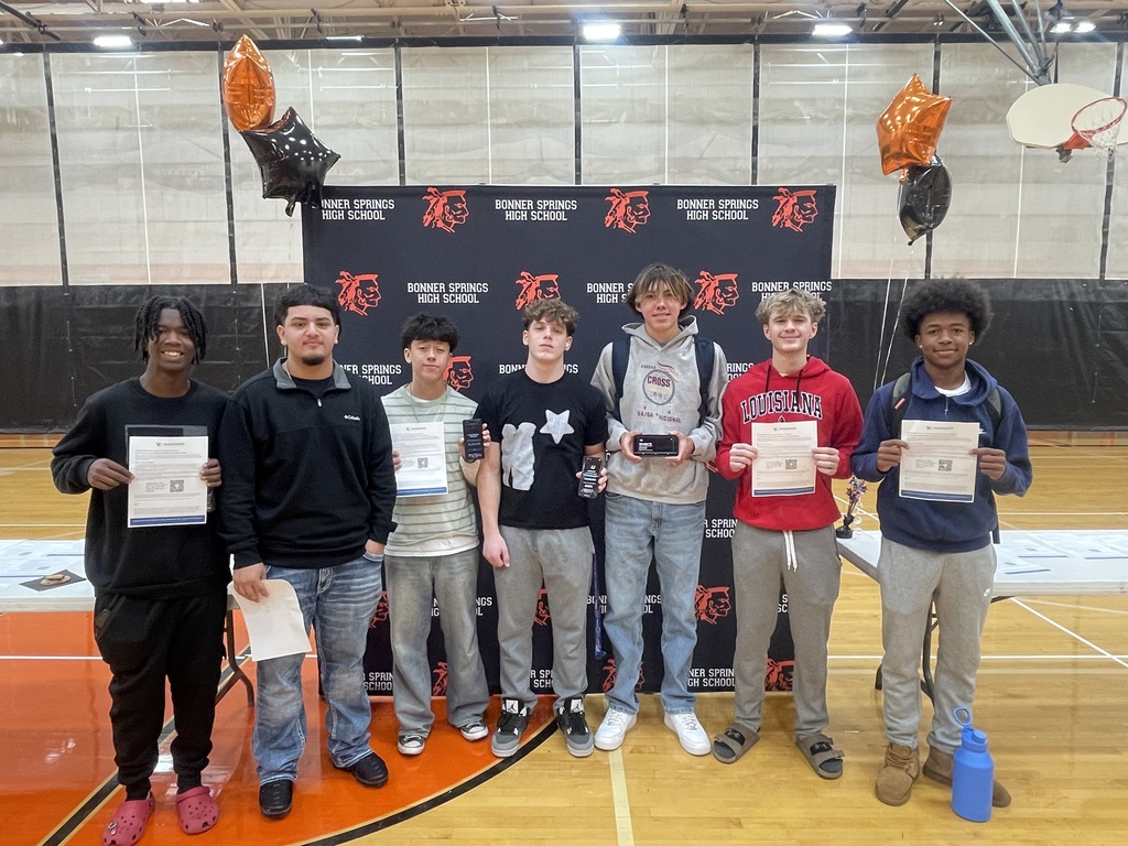 A group of seven students stand side by side in front of a Bonner Springs High School backdrop, each holding up either a scholarship paper or their phone displaying scholarship results. They are smiling and dressed in casual clothing with backpacks.
