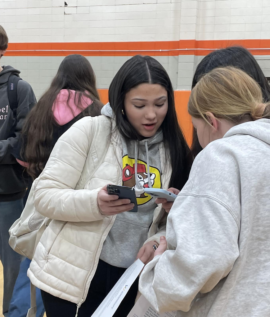 A student wearing a light-colored jacket and hoodie looks down at her phone with an excited, surprised expression while talking with two other students in a gymnasium. She holds papers and a phone as they gather closely, appearing to discuss scholarship results.