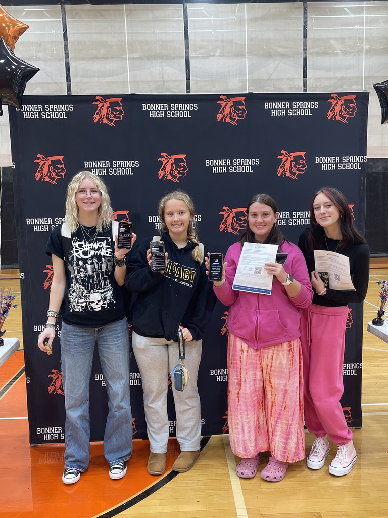 Four students stand in front of a Bonner Springs High School backdrop, smiling and holding up their phones or scholarship papers. They wear casual clothing, including jeans, hoodies, and pink patterned pants.