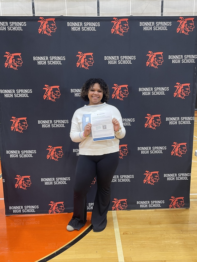 A student stands alone in front of a Bonner Springs High School backdrop, smiling and holding up a printed scholarship notification document.