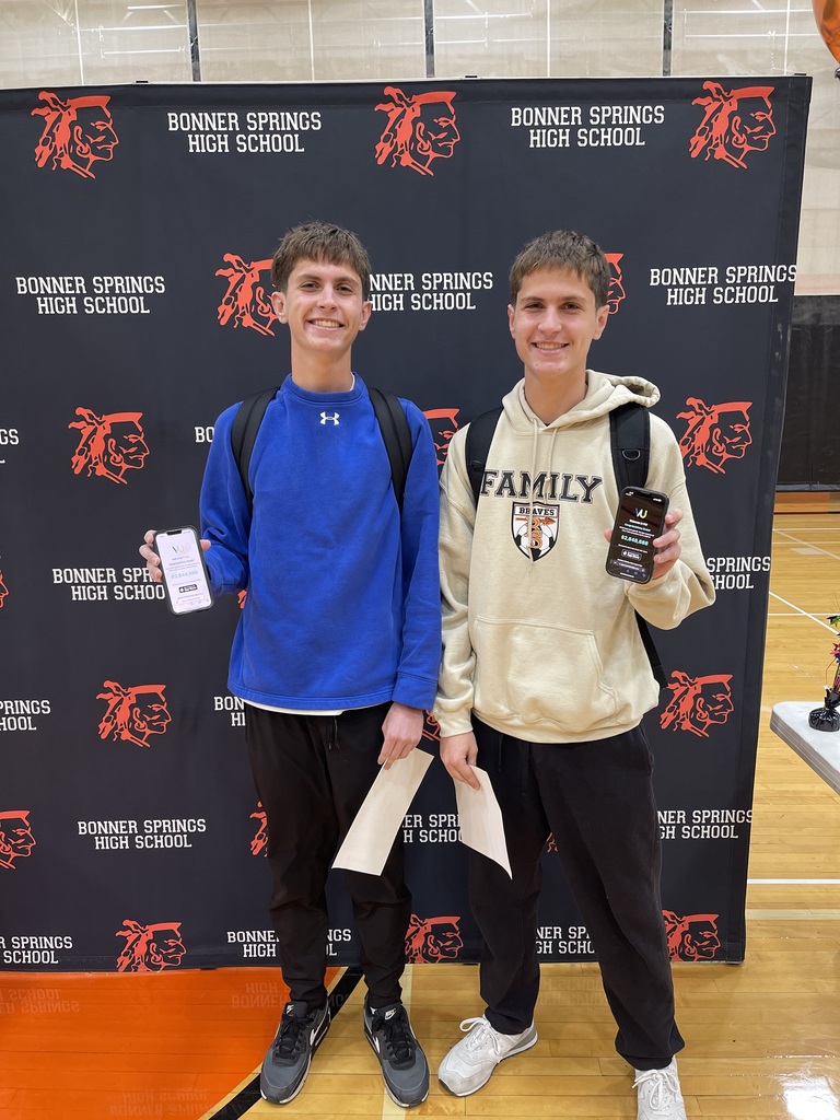 Two students stand in front of a Bonner Springs High School backdrop, smiling and holding up their phones displaying scholarship notifications. Both hold papers in their other hands and wear backpacks.
