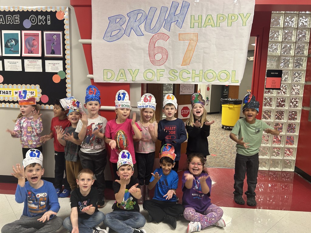A group of kindergarten students wearing handmade “67 Days of School” hats pose and smile in a school hallway. They stand and sit beneath a large colorful banner that reads “BRUH HAPPY 67 DAY OF SCHOOL.” The children have their hands raised playfully toward the camera. Behind them are bright bulletin boards, a red doorway, and a glass-block wall.