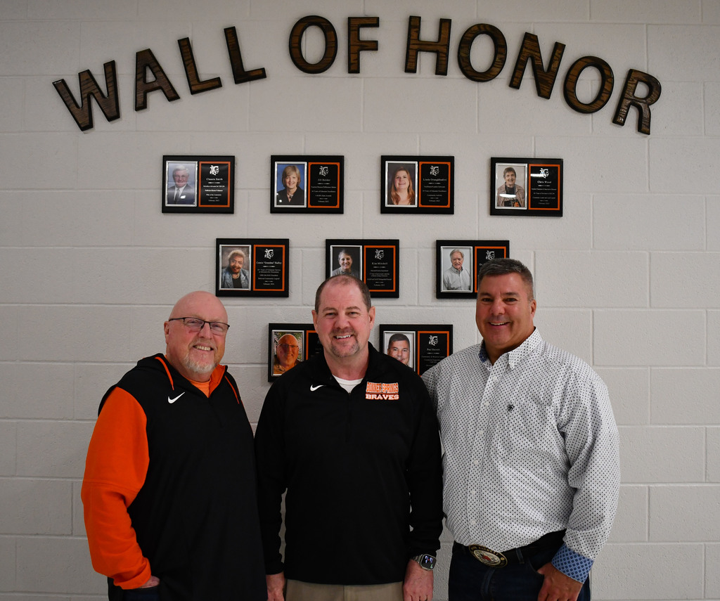 Three men stand smiling in front of a “Wall of Honor” display inside a school building. Behind them is a white brick wall with the words “WALL OF HONOR” in large letters at the top, and a grid of framed plaques featuring photos and names of past honorees. The men are posed side-by-side, with the two on the left wearing black athletic jackets and the man on the right wearing a light-colored button-down shirt.