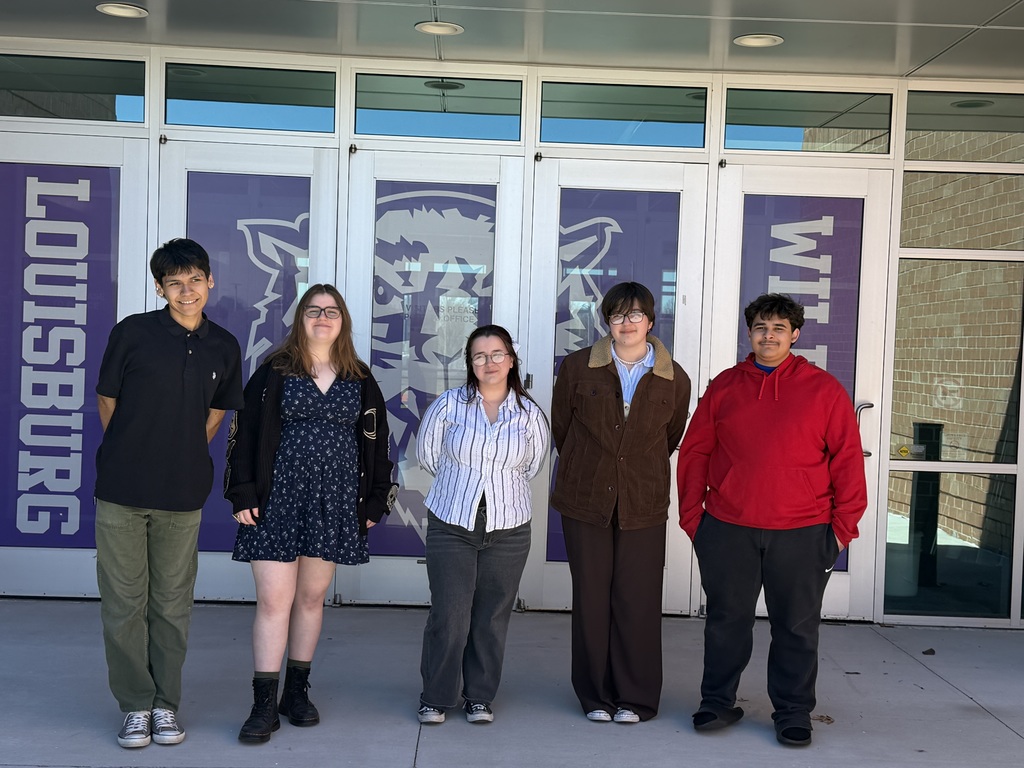 Five people stand in front of a school building, each wearing different clothing. Behind them are large glass doors and large signs.