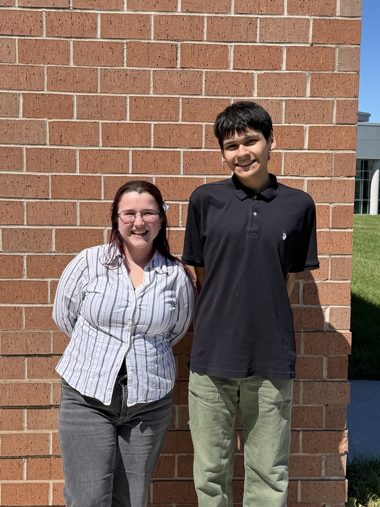 Two individuals stand against a red brick wall; a woman in a white and gray striped shirt, and a boy in black.