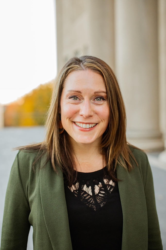 A woman in a green jacket smiles at the camera with autumn trees and a building pillar behind her.