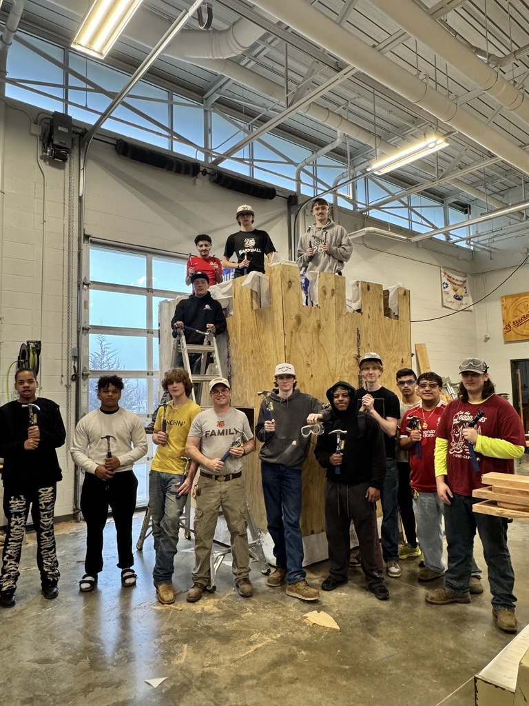 A group of people stand around a wooden structure in a workshop. Some hold tools. The room has windows, ceiling lights, and an open area.