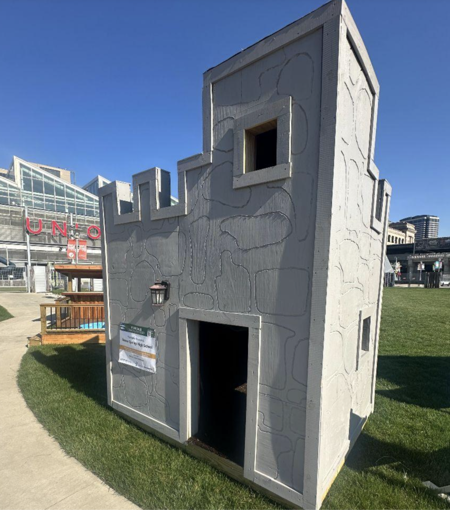 A small castle-like structure stands on grass, featuring a door, windows, and a sign. Behind, buildings and a blue sky are visible.