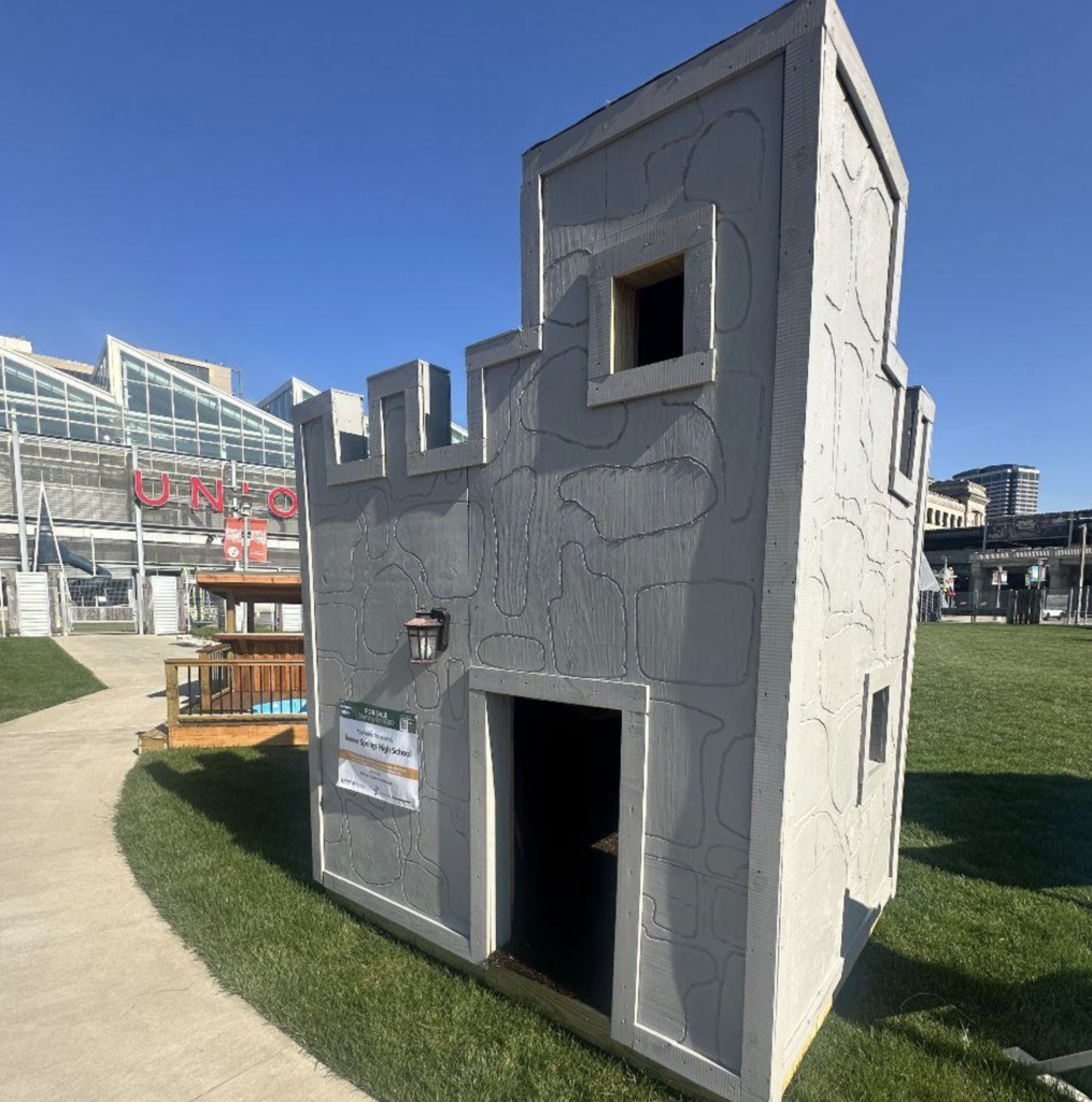 A small, gray, castle-shaped building on a lawn. The building has windows, a door, and a sign.