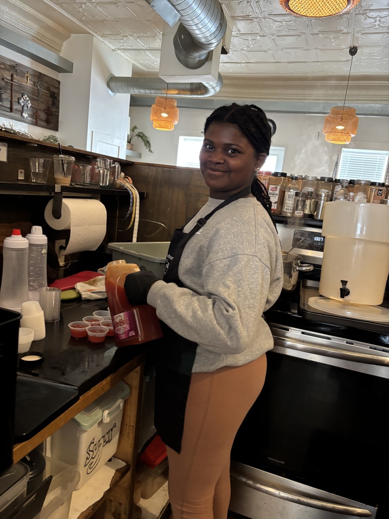 A girl in a gray sweater and apron stands at a kitchen counter with bottles and containers.