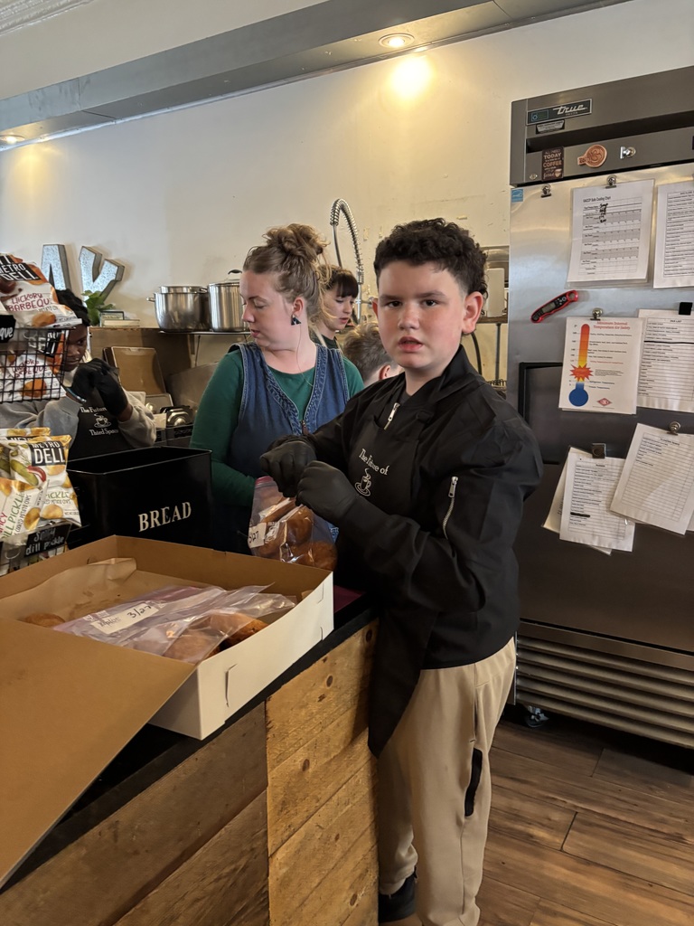 A boy is standing at a counter, holding a box with bread inside. Another person stands behind him.